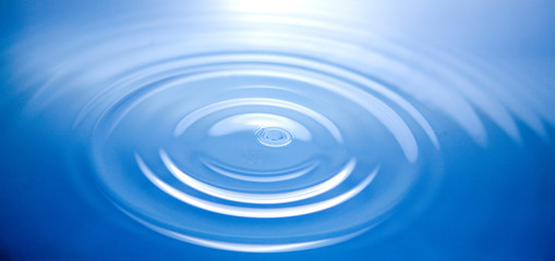 single drop of water hitting the blue surface of water below causing ripples to spread out widely.