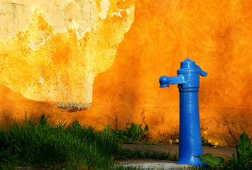 This image features a vibrant blue fire hydrant against an orange textured wall with a large,...