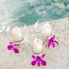 Three candles in glass bowls with purple orchids next to pool.