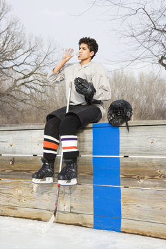 Boy in ice hockey uniform sitting on sidelines drinking water.