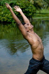 A well-built Asian man practising yoga outside by water