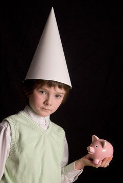 Sad young boy in a dunce cap with a piggybank