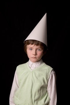 Sad young boy in a dunce cap over a black background