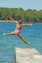 Fototapeta na wymiar Young Girl Jumps Off Dock into Lake Extending Arms and Legs