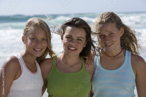 Describe The Three Girls Standing At The Beach "Three teenage girls (13-15) standing on beach, smiling, front view