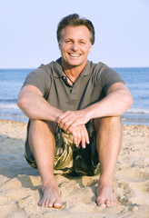 A portrait of a happy 44 year old man sitting on beach.