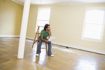 Man sitting on ladder in empty space holding paper thinking