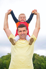 Obraz premium child sits at father on shoulders with lifted hands outdoors