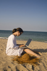 Woman sitting on beach in white dress working on laptop