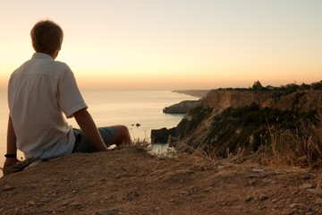 Naklejka na ściany i meble Man sitting on a cliff, staring at a majestic coastline