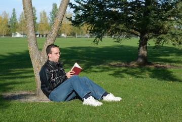 A young man resting by a tree and reading a book