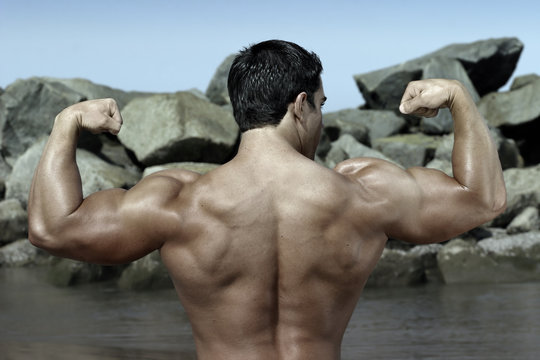 back of a bodybuilder flexing by some rocks on the beach