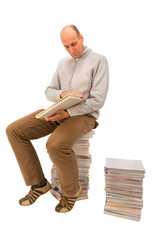 Mann sitzt auf einem Zeitschriftenstapel und schaut ein Buch an