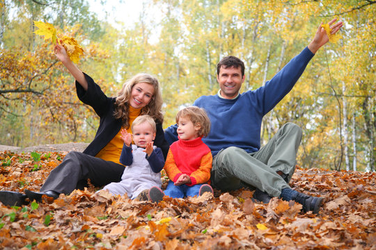 family of four sits in autumn park. parent hands up.