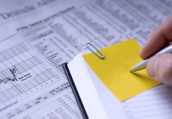 Post It wird im Terminplaner beschriftet. Aktien im Hintergrund.