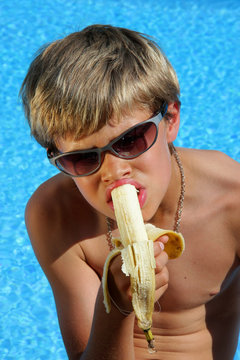 Banane essender Junge am Swimming Pool mit Sonnenbrille