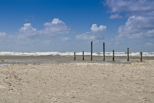 Holzpfähle am Sandstrand vor blauen Himmel mit wolken und wellen der nordsee in St. Peter Ording