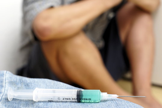 Syringe and sick male addict crouched in background