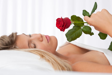 Obraz na płótnie Canvas Beautiful woman lying in white bed holding red rose