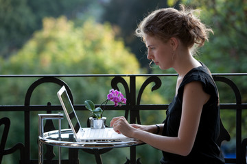 jung Frau schreibt auf einem weißen Laptop auf dem Balkon - 16345784