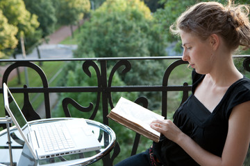 junge blonde Frau sitzt vor Laptop und liest Buch auf Balkon - 16346750
