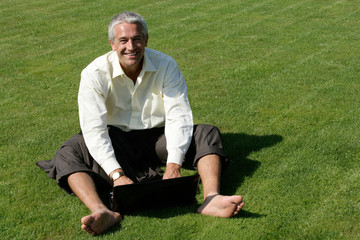 Naklejka na ściany i meble Barefoot businessman sitting on grass and using laptop
