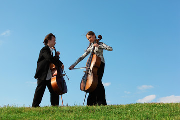 Two violoncellists play on grass against  sky