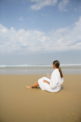 Femme en peignoir assise face à la mer avec une bouteille