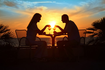 Fototapeta na wymiar Female and man's silhouettes on sunset sit at table with two gla