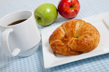 A croissant on a white plate with apples and coffee