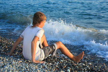 Fototapeta na wymiar sitting teenager boy on stone seacoast, wets feet in water, sitt