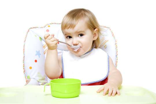 Little girl with bib sitting in baby chair and eating with spoon