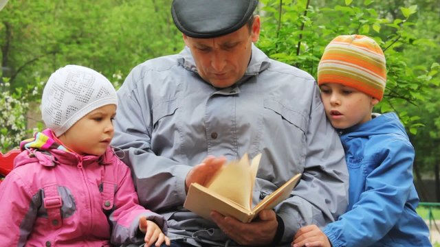 grandfather reads to boys and girls the book in park on a bench