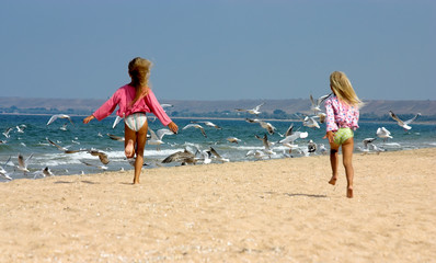 Young girls happily running towards seagulls on seashore