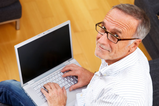mature man with laptop and reading specs looking to camera