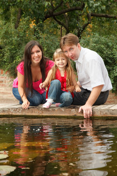 Little girl with parents sits on bank of reservoir in summer