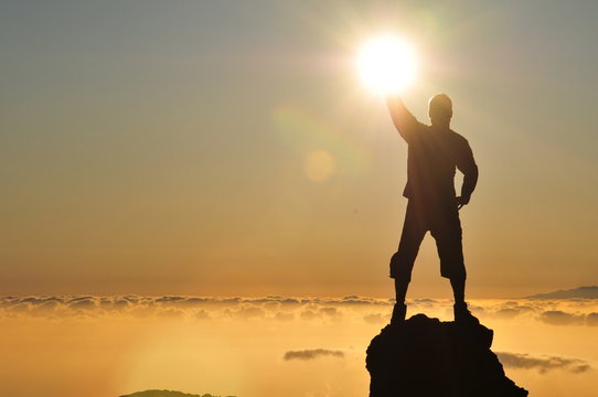 Junger spiritueller Mann auf Berg im Gegenlicht mit Wolken und Sonne. Symbolhaft für einen euphorischen erfolgreichen Held mit viel Energie.