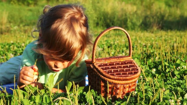 little girl lies in grass near backet, eats cherry and smile