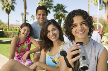 boy (13-15) holding camcorder sitting outdoors with sister (7-9) and parents portrait.