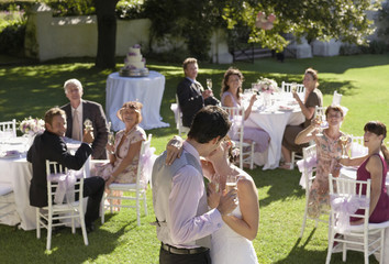 mid adult bride and groom in garden among wedding guests holding wineglasses kissing