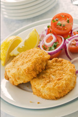 Two fish cakes on a plate with salad and sliced lemons