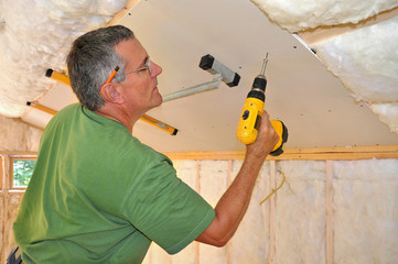 Man using cordless drill to attach drywall panel to ceiling