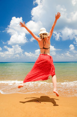 Woman with red sarong and white hat is jumping on the beach