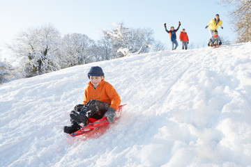 Fototapeta na wymiar Young Boy Sledging Down Hill With Family Watching