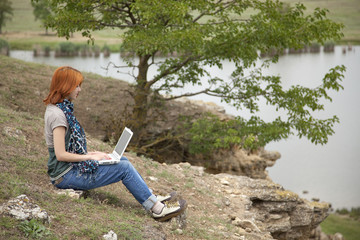 Young beautiful girl with laptop at rock near lake and tree.