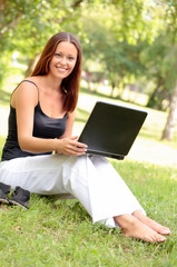 Beautiful young woman sits on a grass in a park with the laptop.