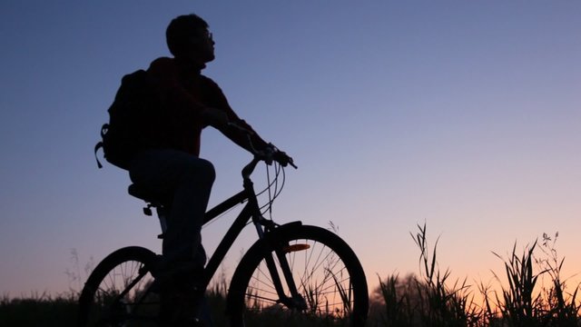 silhouette of man on bicycle stands in field and looks at sky