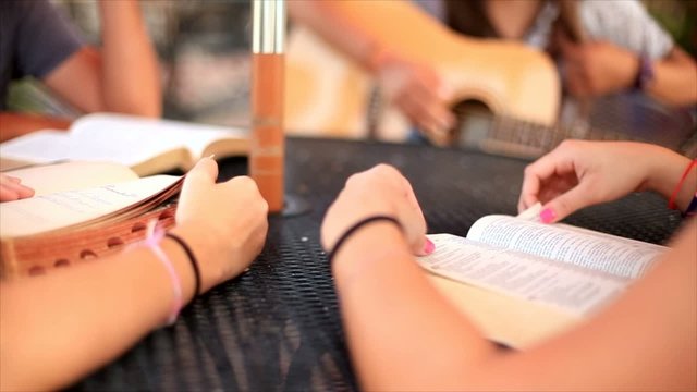 Close up footage of bibles being read by a group of Christian teenagers sitting outside studying and listening to guitar music.