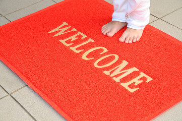 Child Standing On A Red Welcome Mat