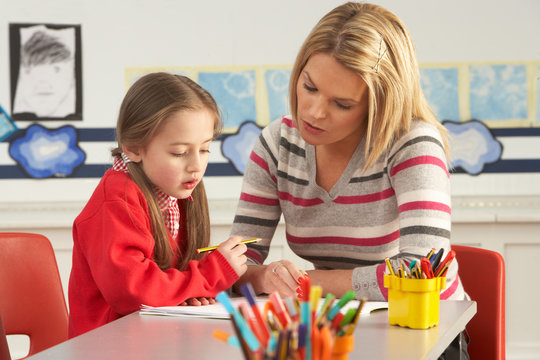 Female Primary School Pupil And Teacher Working At Desk In Class